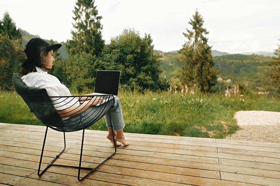 Contact - Woman Sitting in a Patio Chair on a Wooden Deck Overlooking a Beautiful Wooded Area, Using a Laptop